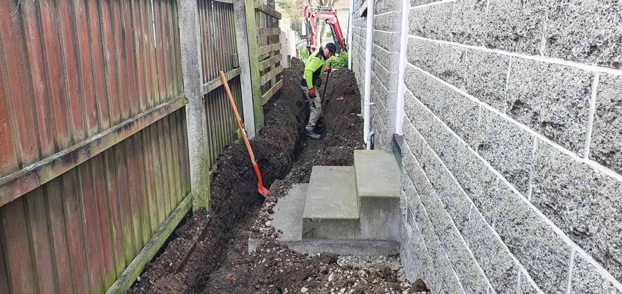 Drainlayer working a narrow trench between a fence and block wall on an earthquake drainage repair in Canterbury